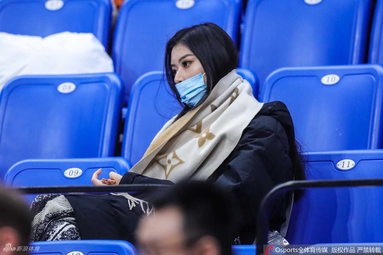 杨诗尹身穿一件设计感十足的白色针织开衫，内搭简约黑色衣物，在看台上低头查看手机。周围是热闹的球迷，而她沉浸在自己的世界里，动静之间更显其独特气质。