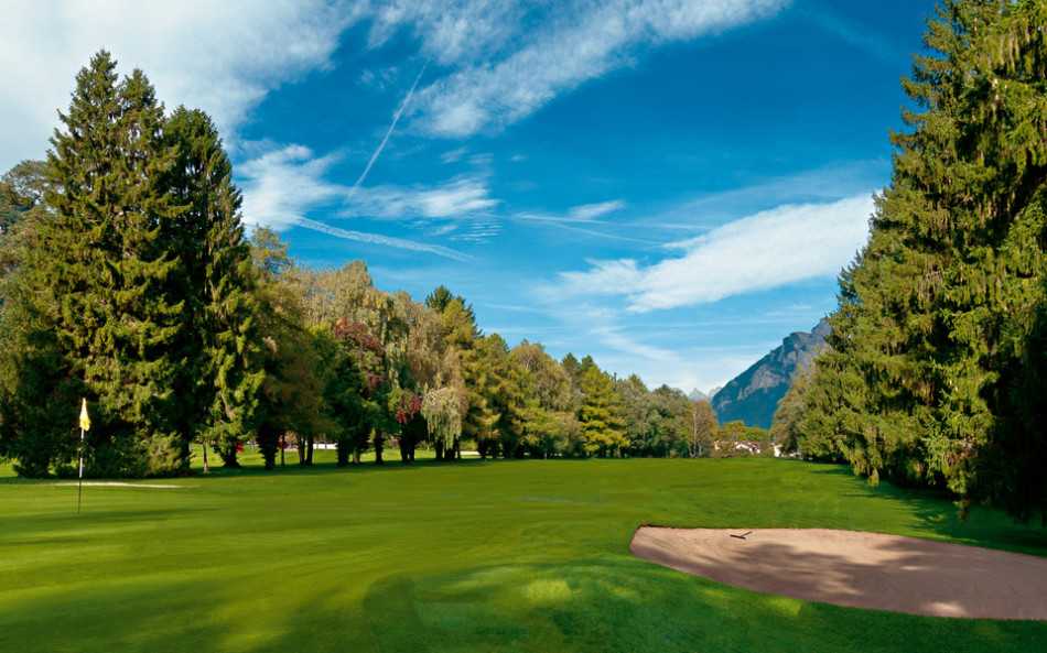 秋日晨曦中，一位高尔夫球手在巴德拉加兹球场优雅开球，金黄的树木与远处雄伟的阿尔卑斯山共同构成一幅宁静的画卷。