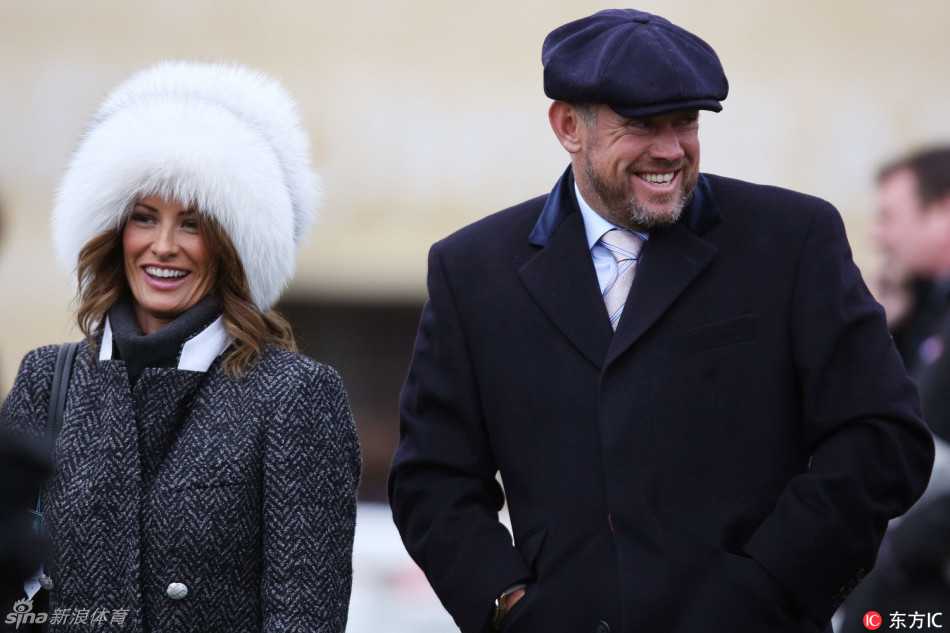 李·维斯特伍德头戴一顶复古风格的贝雷帽，面带标志性的开朗笑容，正与身旁的妻子轻松交谈，背景是熙熙攘攘的切尔滕纳姆赛马节现场。