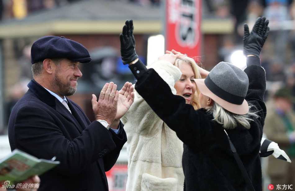 维斯特伍德与妻子并肩而立，两人的目光都投向远方的赛场，神态悠闲，共同享受着切尔滕纳姆赛马节经典的户外社交时光。