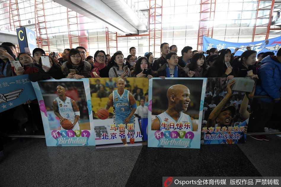 北京队球员孙悦与张庆鹏并肩走出机场，孙悦面带标志性微笑挥手致谢，张庆鹏则神情轻松，两人身上背着运动背包，身旁围绕着一路小跑跟拍的狂热球迷。