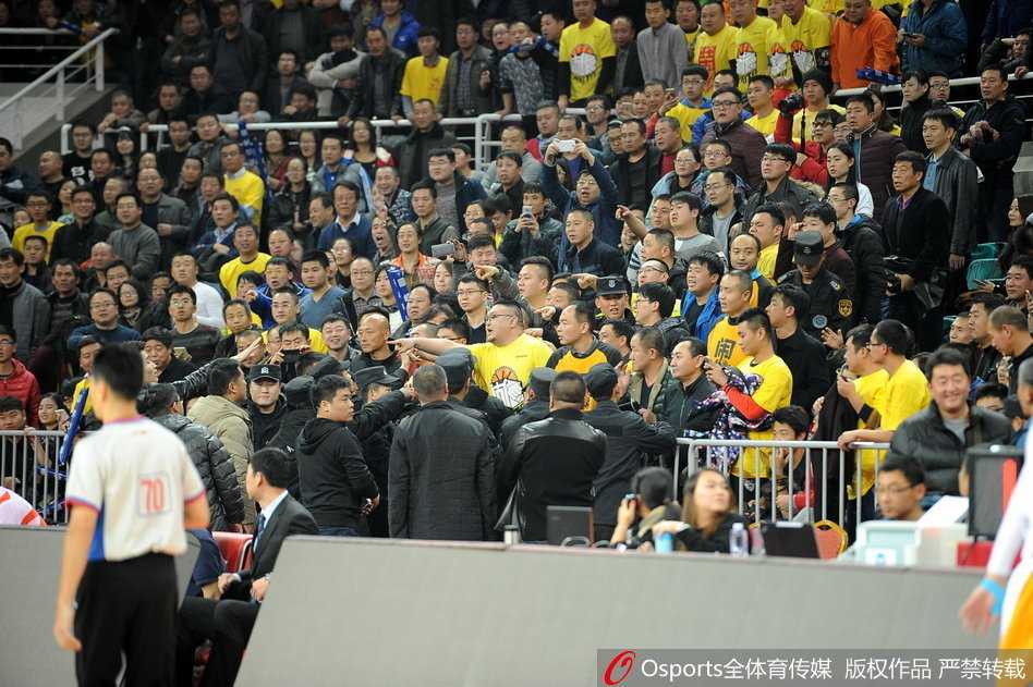 看台上的球迷们集体站立，目光齐刷刷地投向场地一角的冲突区域，许多人高举着双臂，面部表情因高声呼喊而变得扭曲，现场气氛紧张到了极点。