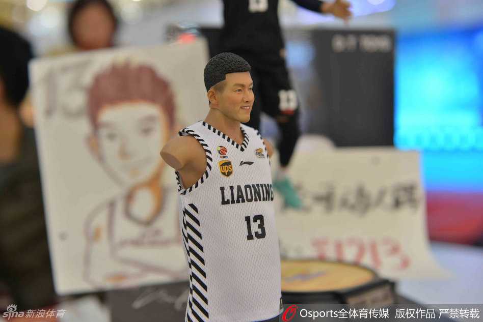 一位身穿黑色外套的美女双手捧着郭艾伦人偶，将人偶举在脸颊旁，闭眼微笑做出亲吻的姿势，身后的背景板印着郭艾伦巨大的赛场英姿。