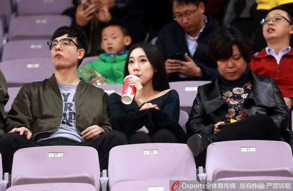 戴眼镜的知性美女球迷置身于热闹看台，她面带温婉微笑，在人群中显得格外沉静，仿佛正沉浸于比赛的紧张节奏中。