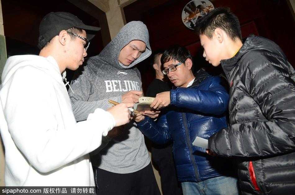 翟晓川身穿蓝色休闲装，面带阳光微笑低头为小球迷签名，周围人群举起手机记录这追星时刻