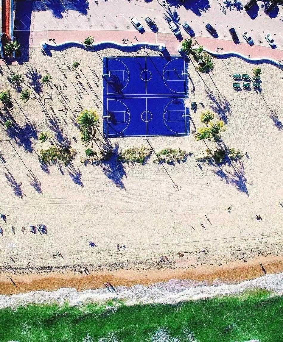 夕阳余晖下，从高处俯瞰篮球场，暖色调的灯光与蓝色地面形成鲜明对比，球员的影子被拉长，如同舞台上的舞者。