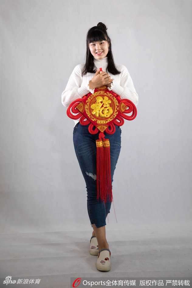 南粤玫瑰绽新春:广东女篮的荣耀与传承 黄思静手持一张精致的金色福字卡,对着镜头俏皮地眨眼,展现出赛场之外活泼可爱的一面,背景中隐约可见其他队友正在相互整理着装,气氛温馨和谐。