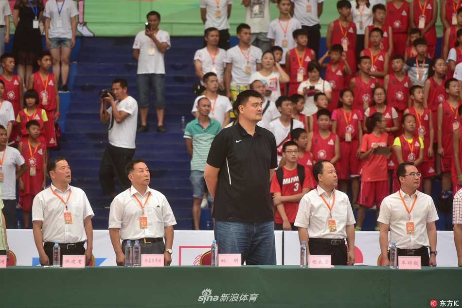 姚明身穿白色衬衫在场边临场指挥，他微微弯腰，右手比划着战术手势，身边围着一群仰着头认真听讲的篮球少年，仿佛一棵参天大树守护着一片新苗