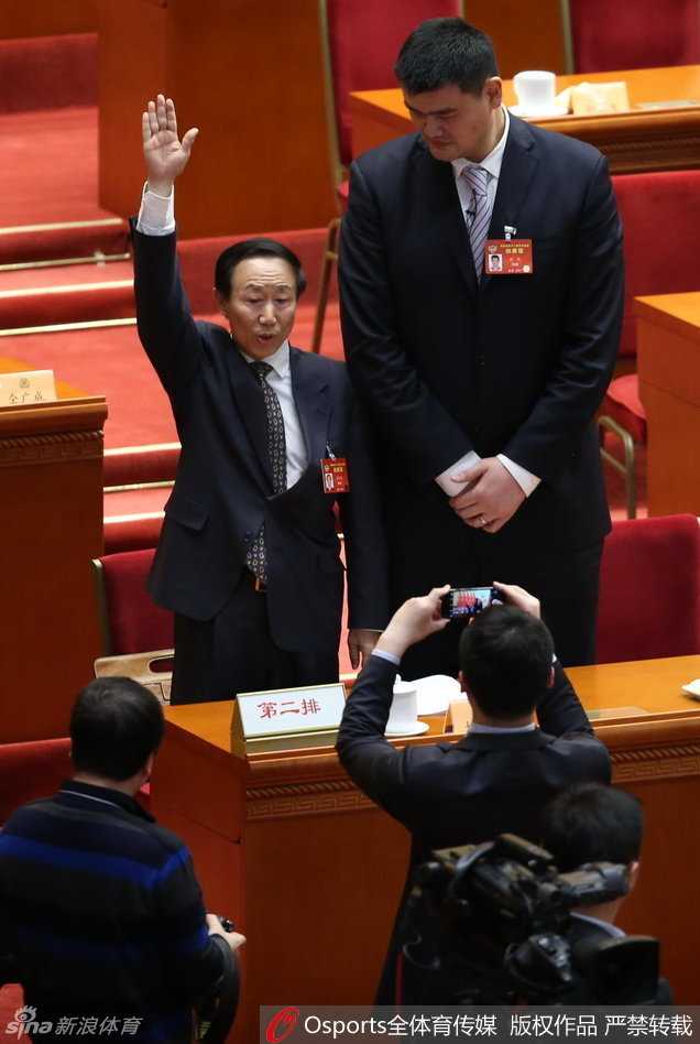 姚明站在人民大会堂的会议厅内，正装笔挺，面带从容的微笑，标志性的身高让他即便在人群中也能被一眼认出，背景是庄严肃穆的会场环境，记录下这位体育人履职的珍贵瞬间。