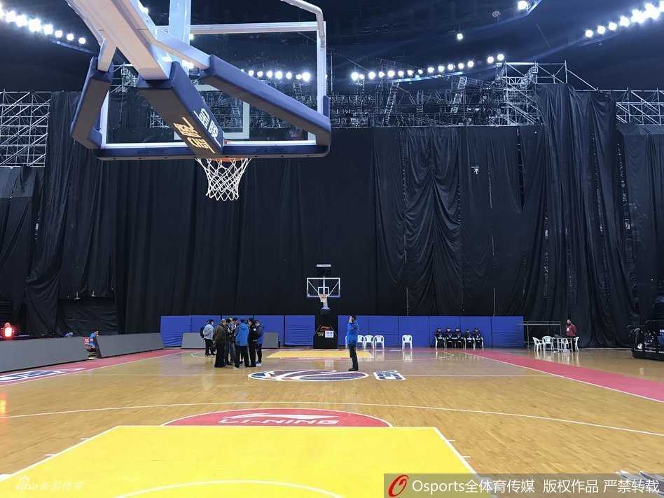 几名身穿工作服的工人正在舞台的脚手架旁进行最后的检查，他们身后是被黑布严密遮盖的舞台主体，背景里青岛主场的冠军旗和退役球衣横幅若隐若现，现实与荣誉在此刻形成鲜明对比。