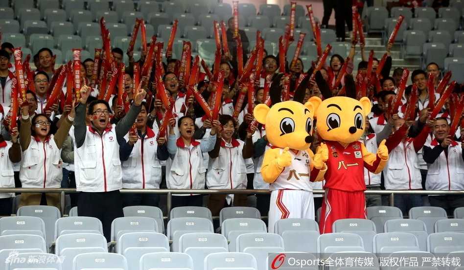 火箭精神燃情看台:八一男篮吉祥物与球迷的难忘互动 吉祥物站在看台台阶上向二楼的观众抛洒小礼物,它仰着头用力挥舞着手中的纪念品,灯光照亮了它身上红色的球衣,上方无数双手臂高高举起,仿佛一片欢呼的森林