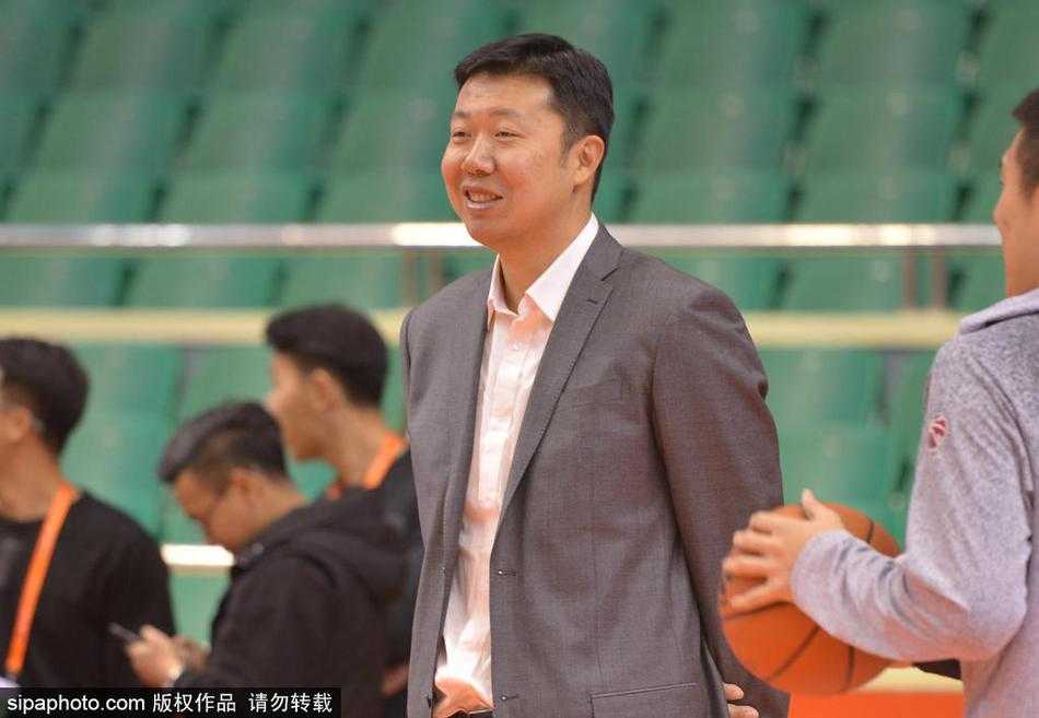 王治郅身穿黑色羽绒服站在赛场中央，双手插兜，面带微笑地注视着远处正在热身的八一队球员，眼神中充满了鼓励与期待。