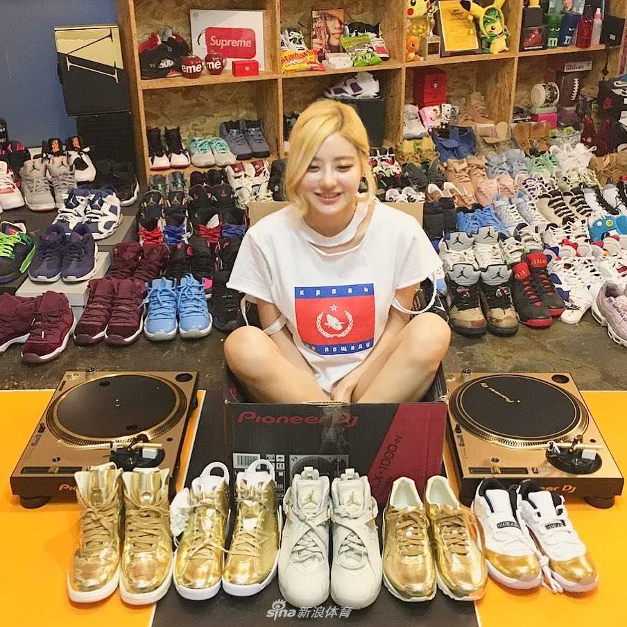 DJ Soda 半蹲在地上，身后是堆积如山的Nike与Jordan鞋盒，她穿着一袭白色连衣裙，金色的短发显得格外俏皮，眼神中透露出对自己收藏帝国的满足与骄傲。