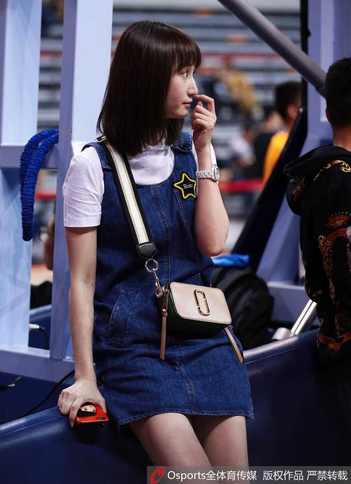 刘美在赛场边低头查看手机，嘴角带着一抹温柔的笑意，牛仔裙的简约设计衬托出她清新脱俗的气质，与身后紧张的比赛氛围形成有趣对比。