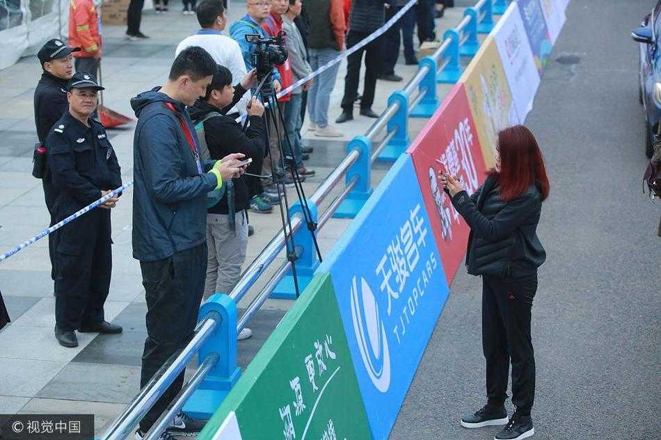 镜头拉远，冬日娜和杜锋并肩站在赛道旁的护栏边，周围是攒动的人头和飘扬的旗帜。完成扫码的两人似乎正进行着简短的交流，身后巨大的赛事背景板无声地宣告着这场亚洲田径盛事的主场。