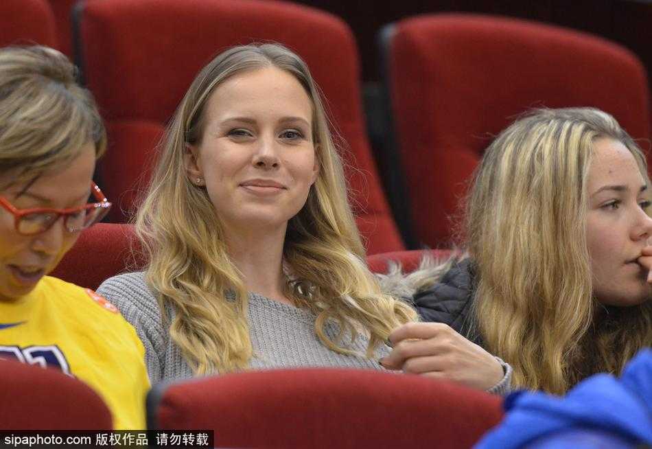 莫泰妻子在看台上与身边友人交谈，笑容温婉大气，金色的长发在灯光下熠熠生辉，尽显东欧女性的独特魅力