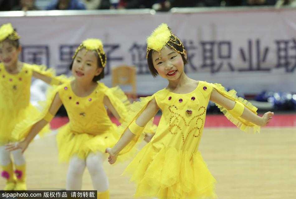 萌娃空降甬城赛场 CBA迎来最萌啦啦队 几名小队员站成一排,手里拿着色彩鲜艳的花球,背景是巨大的CBA赛场广告牌。她们在等待退场,眼神中带着好奇与一丝疲惫,但依然紧紧抱着花球,舍不得放下。