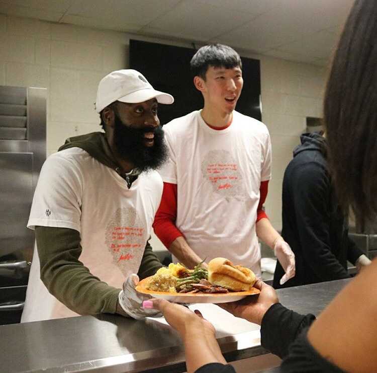 在摆满餐盘的桌子前，哈登露出标志性的大胡子笑容，耐心地为民众服务，周琦在一旁协助，画面充满了社区大家庭的温馨感。