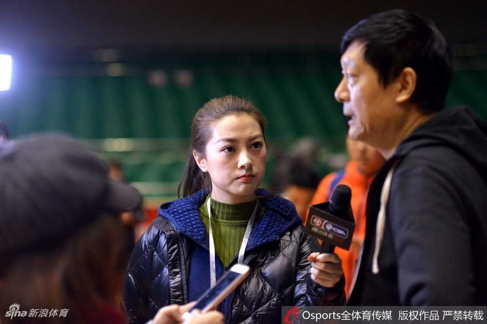 赛场背后探营记:美女记者直击新疆广汇与山东高速男篮高强度备战 全景镜头下,山东男篮的队员们正在进行快攻上篮练习,篮球在空中划出弧线。美女记者站在场边的摄影位,与身后巨大的球队标志相映成趣,展现出职业体育报道的规范与热情。