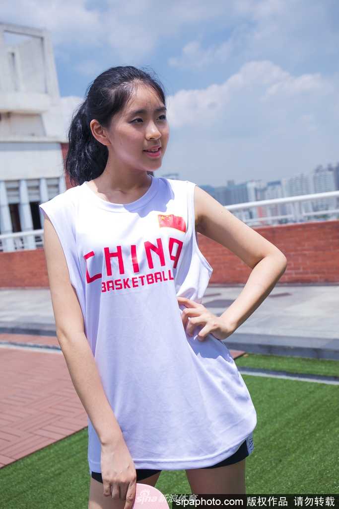 女孩身穿白色运动背心搭配牛仔短裤，健康的小麦色肌肤在阳光下泛着光泽，她单手叉腰，身姿挺拔，展现出篮球宝贝特有的自信与健美。