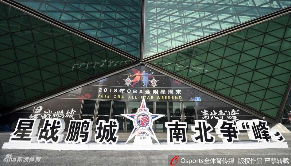 深圳龙岗大运中心在湛蓝天空下展现出独特的六边形建筑结构，银灰色的外墙熠熠生辉，广场上旗帜迎风招展，迎接着八方球迷的到来。